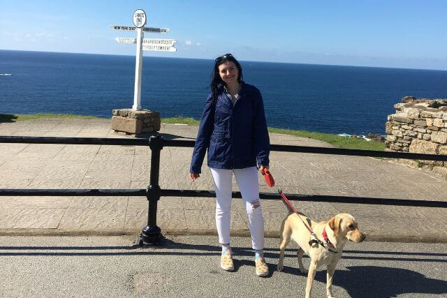 A woman stands with a dog in front of the Land's End sign with the sea in the background.