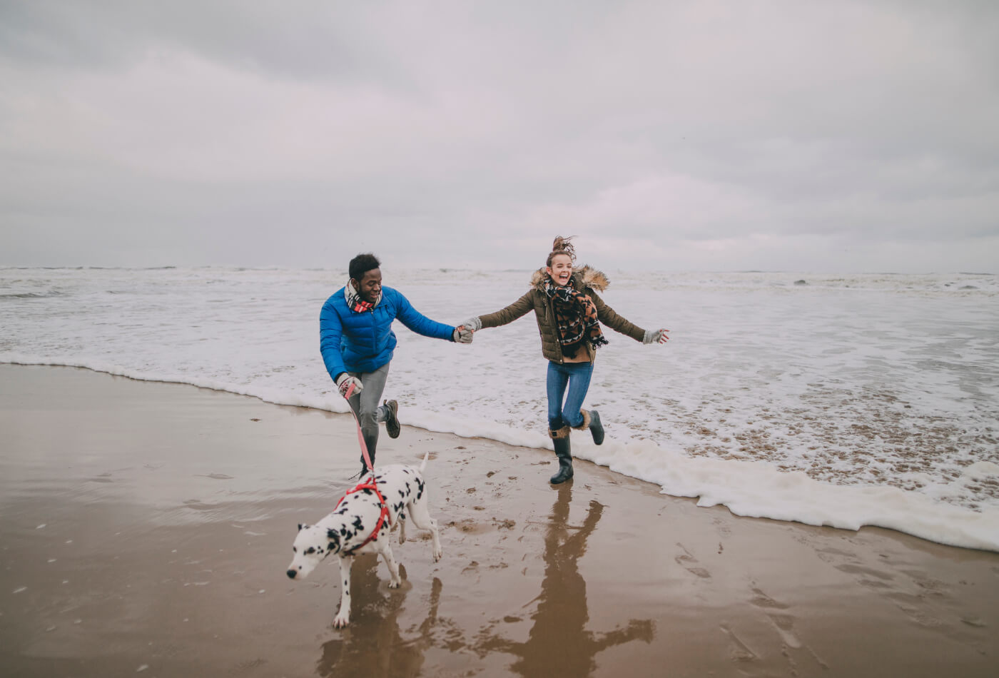 Couple running on beach in Cornwall in winter with Dalmation dog.