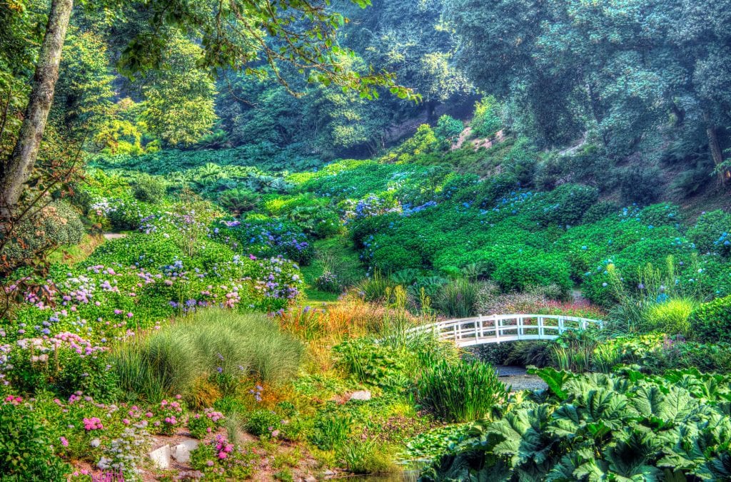 Gardens in a valley with flowers and shrubbery with a white draw bridge over a stream.