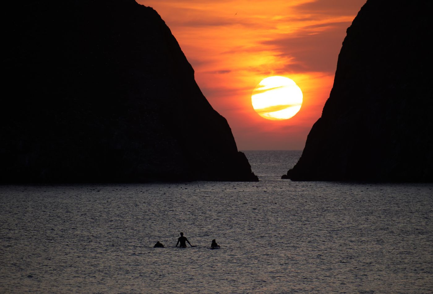 Three surfers sit and watch the sunset in Holywell Bay as it sets behind the clouds and the v shape rock formation.